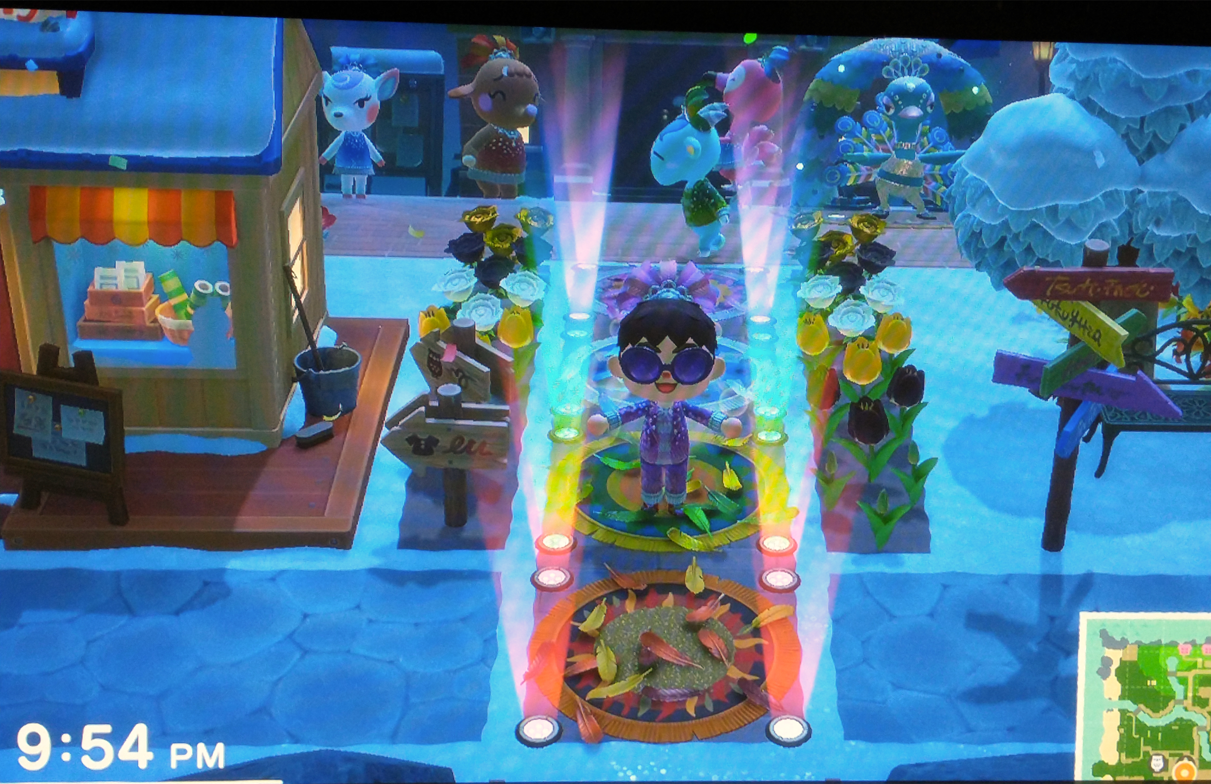 A screenshot from the video game Animal Crossing: New Horizons. A cartoon avatar stands in the middle of a rainbow-colored pathway, wearing a sparkly suit and ostentatious sunglasses, gesturing as if to welcome the viewer down the path.